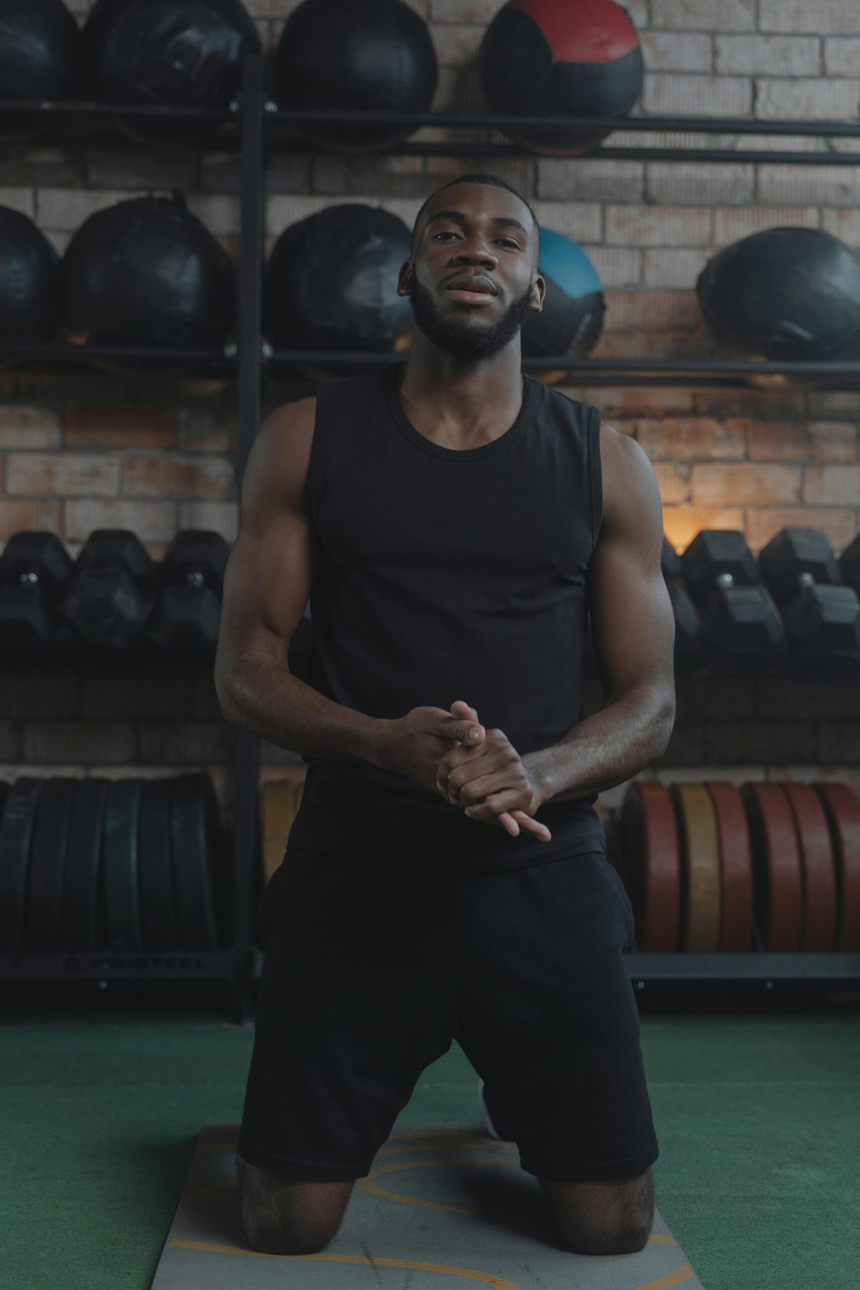 Athletic man kneeling indoors in a gym setting surrounded by fitness equipment.